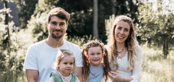 Familie May sitzt lächelnd auf einem Baumstamm in einer sonnigen Wiese – natürlicher, fröhlicher Moment im Freien